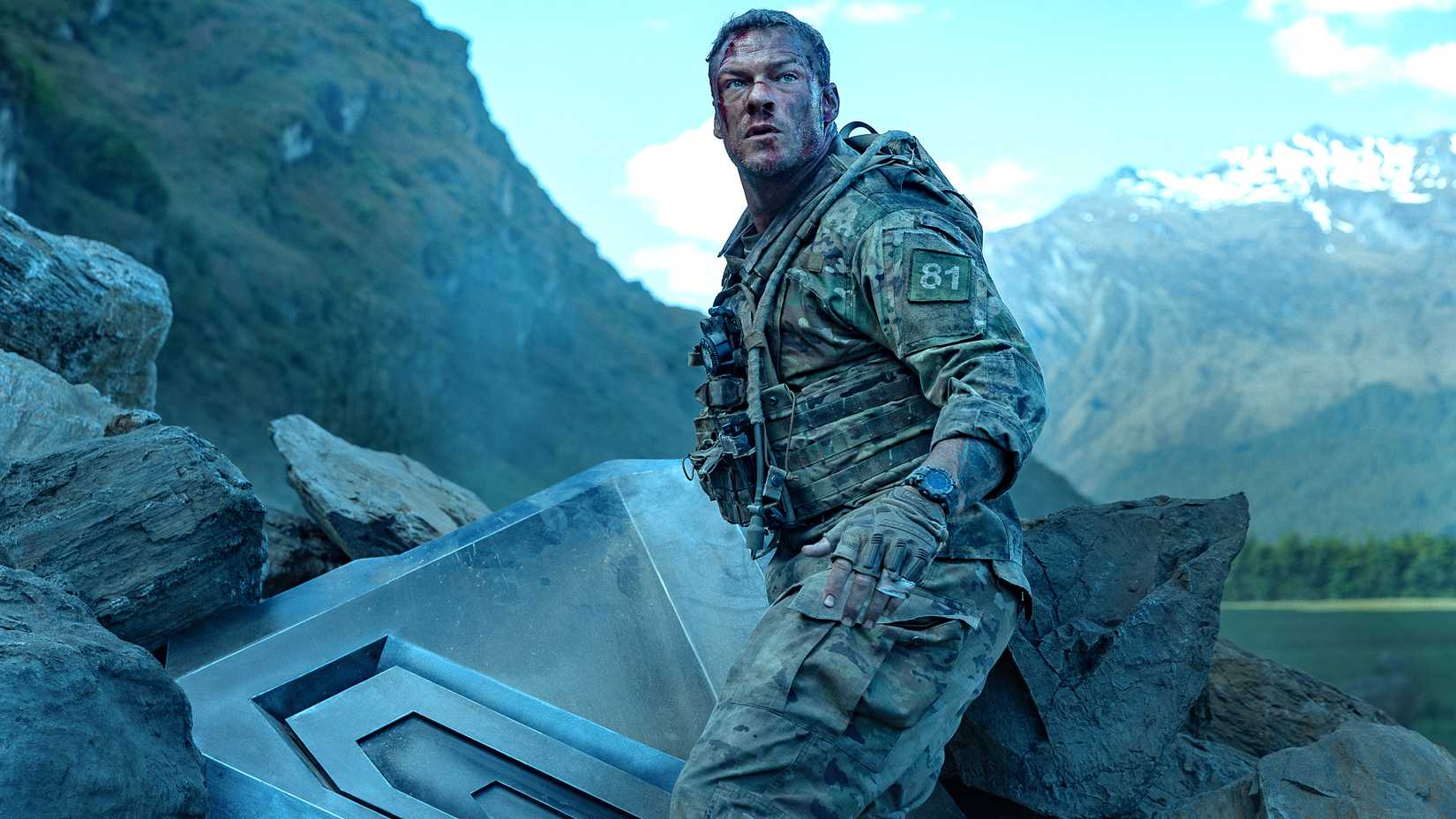 Alan Ritchson, caracterizado como o personagem 81, ao lado de destroços de máquina em cena de War Machine